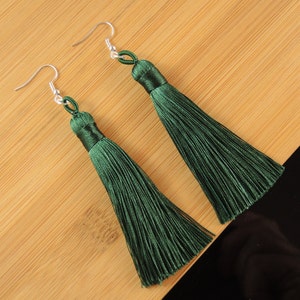 May include: A pair of long, dark green tassel earrings with silver hooks. The silky tassels are displayed on a light brown wooden surface. The earrings are a stylish accessory.