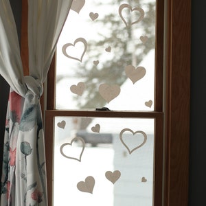 May include: A window with white paper hearts cutouts on the glass. The window is framed in wood and has a white curtain on the left side.