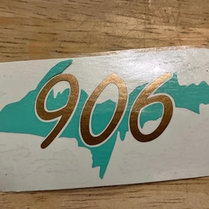 May include: A rectangular sticker featuring the number "906" in a metallic gold color. The numbers are superimposed over a teal-colored outline of a landmass. The sticker has rounded corners and is set against a wooden surface.