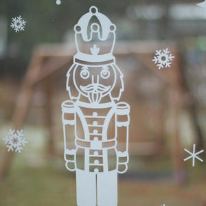May include: White silhouette of a nutcracker with a crown on a frosted window with snowflakes.