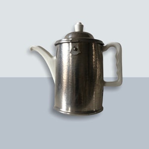 May include: A silver coffee pot with a white lid and handle. The pot has a hammered metal finish.