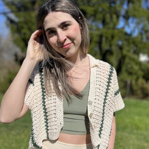 Puede incluir: Un top de crochet blanco con rayas verdes y un cierre de botón único. El top es corto y tiene mangas cortas.