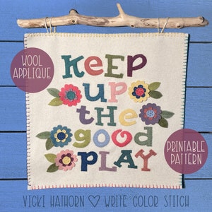 May include: A square, off-white wool applique with the words "KEEP UP THE GOOD PLAY" in colorful, stitched letters. Decorative felt flowers are scattered around the text. The piece is suspended from a wooden branch.