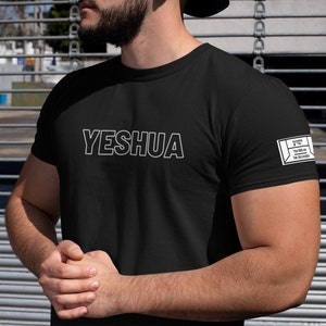 Pode incluir: Camiseta preta com texto branco que diz "YESHUA" e um remendo retangular branco na manga direita com texto em hebraico e as palavras "YESHUA IS THE DELIVERER".