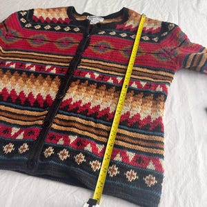 May include: A button-down cardigan sweater with a geometric pattern in red, black, tan, and blue. The sweater has a round neckline and long sleeves. A yellow measuring tape is laid across the sweater, showing the length in inches.