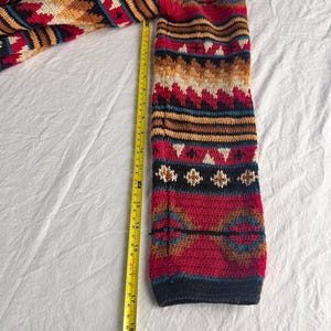 May include: A colorful, patterned sleeve of a knitted garment, featuring a mix of red, blue, tan, and white geometric designs. A yellow measuring tape shows the sleeve is approximately 18 inches long.
