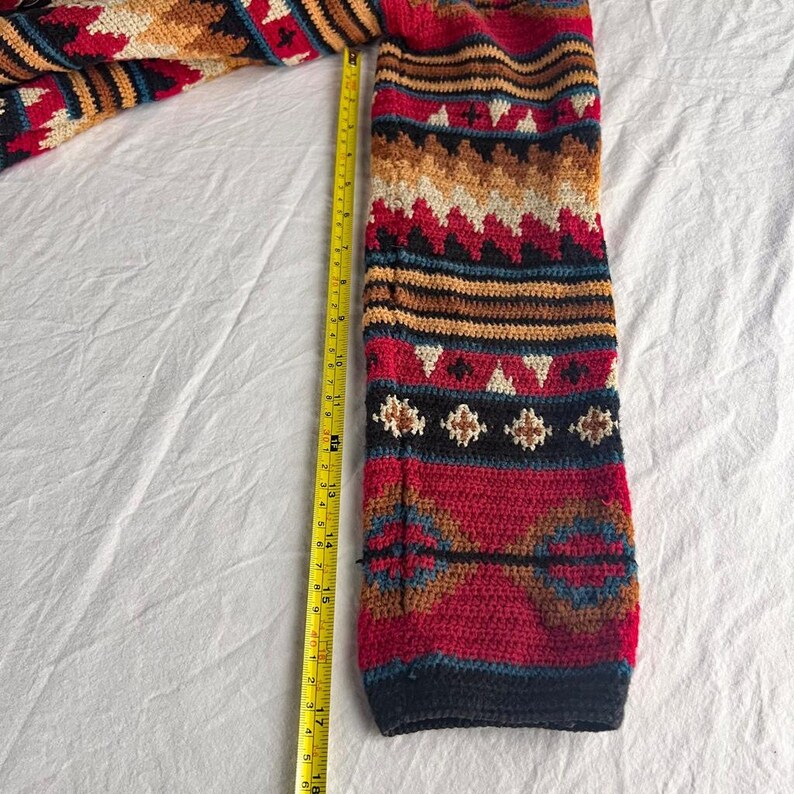 May include: A colorful, patterned sleeve of a knitted garment, featuring a mix of red, blue, tan, and white geometric designs. A yellow measuring tape shows the sleeve is approximately 18 inches long.