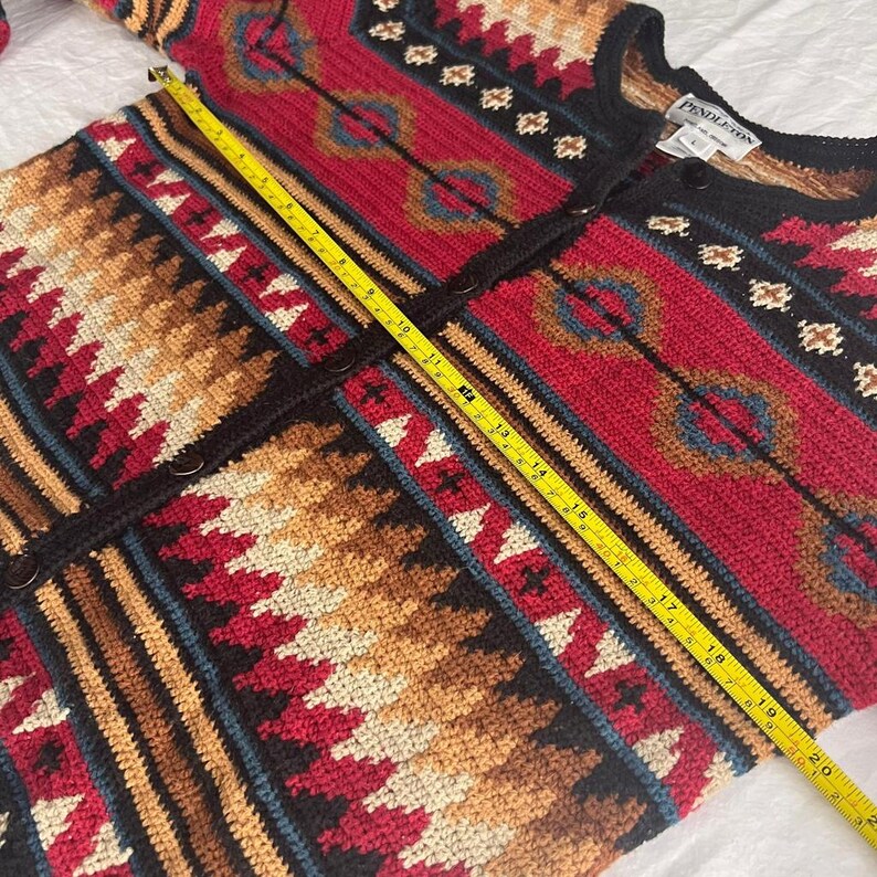 May include: A colorful, patterned cardigan sweater with a button-up front. The sweater features geometric designs in red, brown, black, and blue. A yellow measuring tape is laid across the sweater, showing a measurement of approximately 20 inches.