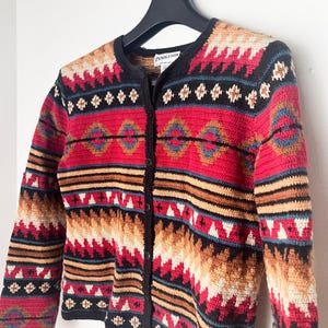 May include: A button-down cardigan sweater with a Southwestern pattern. The sweater features a black collar and trim, with a red, tan, black, and blue geometric design. The label reads "Pendleton".