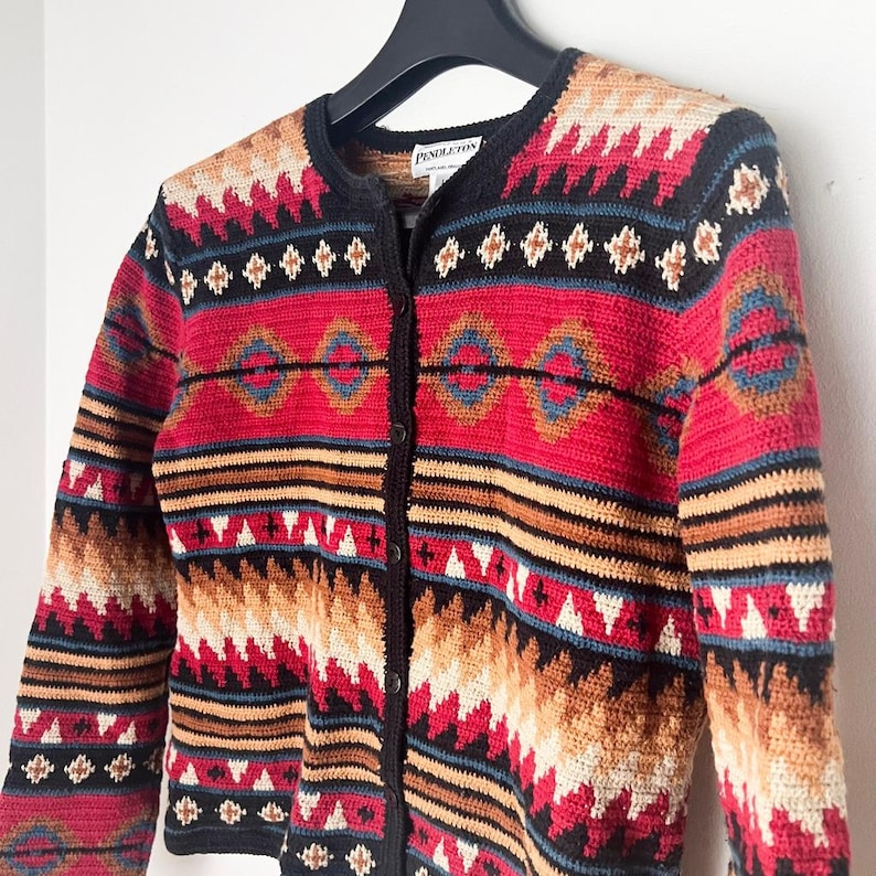 May include: A button-down cardigan sweater with a Southwestern pattern. The sweater features a black collar and trim, with a red, tan, black, and blue geometric design. The label reads "Pendleton".