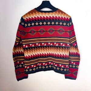 May include: A colorful, patterned sweater with a black collar and cuffs. The sweater features a geometric design in red, white, brown, and blue. The garment is displayed on a black hanger against a white background.