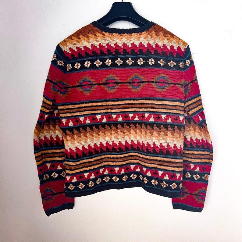 May include: A colorful, patterned sweater with a black collar and cuffs. The sweater features a geometric design in red, white, brown, and blue. The garment is displayed on a black hanger against a white background.