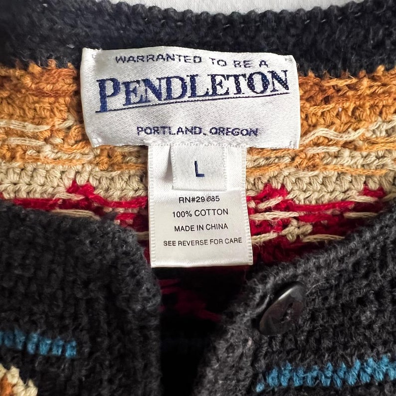 May include: Close-up of a Pendleton cardigan label. The label reads "WARRANTED TO BE A PENDLETON PORTLAND, OREGON" and includes size "L" and "100% COTTON." The sweater has a button closure and a colorful knit pattern.