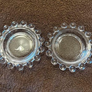 May include: Two clear glass dishes with a circular design. Each dish has a central bowl shape surrounded by a ring of small, clear, spherical accents. The dishes are displayed on a brown surface.