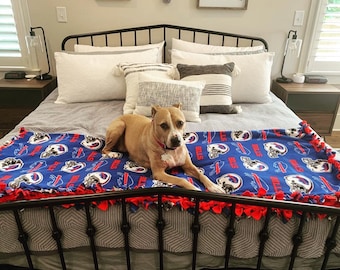 May include: A brown and white dog laying on a blue and red fleece blanket with the word "Bills" repeated on the blanket. The dog is laying on a bed with white pillows and a black metal frame.