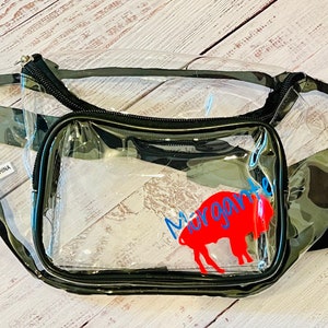 May include: Clear plastic fanny pack with a camouflage pattern. The pack has a zippered pocket with a red buffalo silhouette and the text "Margarita" in blue.