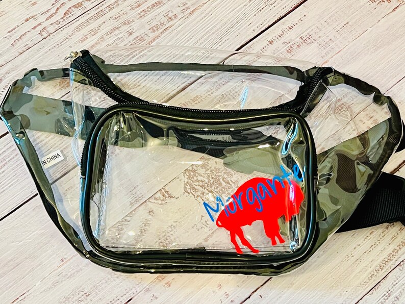 May include: Clear plastic fanny pack with a camouflage pattern. The pack has a zippered pocket with a red buffalo silhouette and the text "Margarita" in blue.