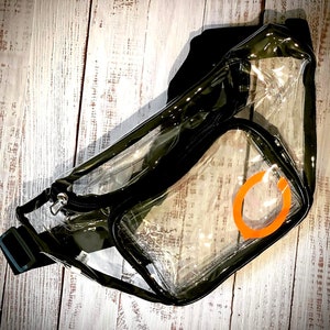 May include: Clear plastic fanny pack with a black adjustable strap and a zipper closure. The pack has a large clear pocket with an orange circle design.