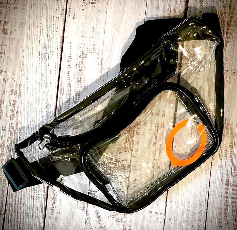May include: Clear plastic fanny pack with a black adjustable strap and a zipper closure. The pack has a large clear pocket with an orange circle design.