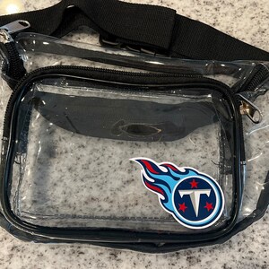 May include: Clear plastic fanny pack with a black adjustable strap and a zipper closure. The pack has a blue and red Tennessee Titans logo sticker on the front.