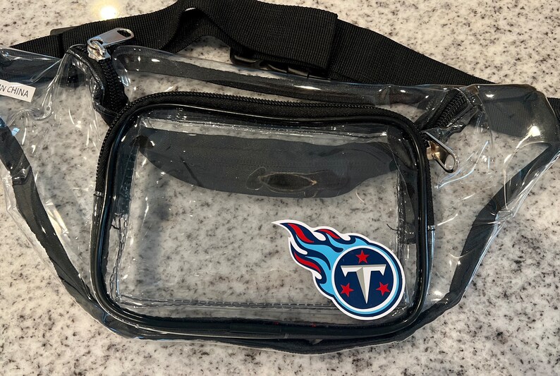 May include: Clear plastic fanny pack with a black adjustable strap and a zipper closure. The pack has a blue and red Tennessee Titans logo sticker on the front.