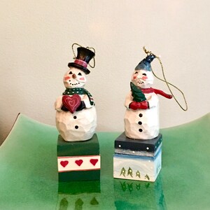 Op de afbeelding: Twee houten sneeuwman ornamenten met beschilderde details. De sneeuwman links houdt een rood hart vast en draagt een groene sjaal. De sneeuwman rechts houdt een groene boom vast en draagt een rode sjaal. Beide sneeuwmannen staan op houten bases met beschilderde details.