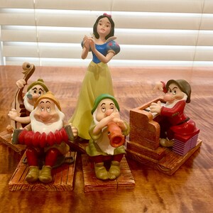 Op de afbeelding: Sneeuwwitje-figuur met een gele jurk en een rode strik in haar haar, staand voor vier dwergfiguren die muziekinstrumenten bespelen. De dwergen dragen kleurrijke kleding en zitten op houten platforms.