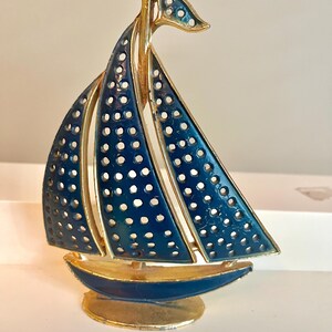 May include: A decorative sailboat figurine with a gold-tone frame and navy blue enamel sails. The sails have a perforated design. The boat sits on a gold base and has a gold mast and flag.