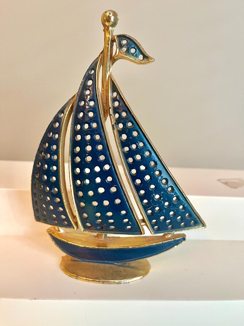 May include: A decorative sailboat figurine with a gold-tone frame and navy blue enamel sails. The sails have a perforated design. The boat sits on a gold base and has a gold mast and flag.