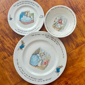 May include: A set of three white ceramic dishes featuring illustrations and text from a children's story. The set includes two plates and a bowl, each with a different scene and text around the rim. The illustrations depict rabbits in various settings.