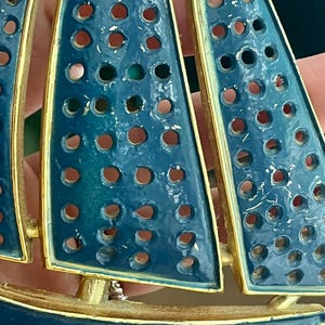 May include: A vintage sailboat brooch with blue enamel sails and a gold-tone metal frame. The sails have a series of circular cutouts. The brooch is a nautical-themed accessory.