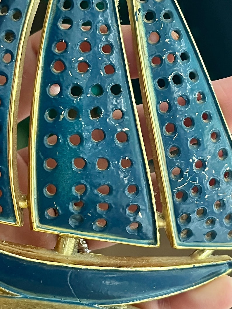 May include: A vintage sailboat brooch with blue enamel sails and a gold-tone metal frame. The sails have a series of circular cutouts. The brooch is a nautical-themed accessory.