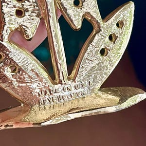 May include: Close-up of a gold-toned metal brooch with a floral design. The brooch has a textured surface and several small holes. The text "MADE IN USA" and "BEN-NERICO" is visible on the brooch.