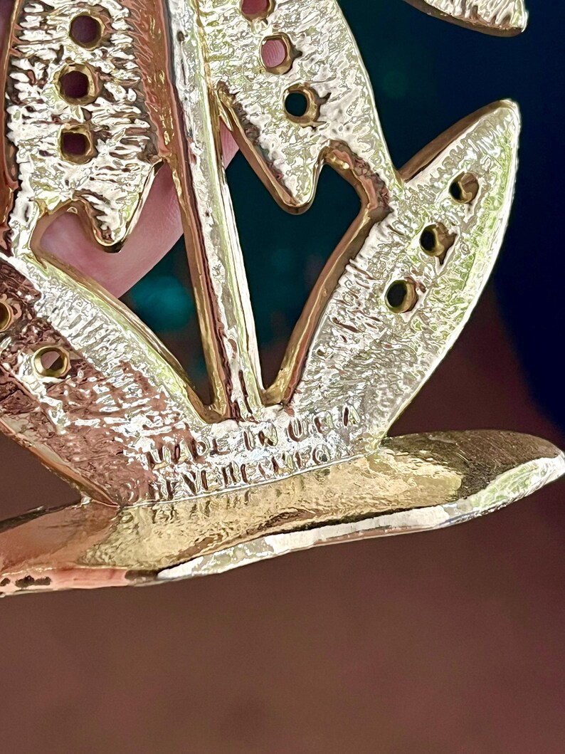 May include: Close-up of a gold-toned metal brooch with a floral design. The brooch has a textured surface and several small holes. The text "MADE IN USA" and "BEN-NERICO" is visible on the brooch.