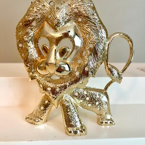 May include: A gold-colored lion figurine with a detailed mane and tail. The lion is standing on all fours, with a textured surface and small holes throughout. The figurine has a regal and decorative appearance.
