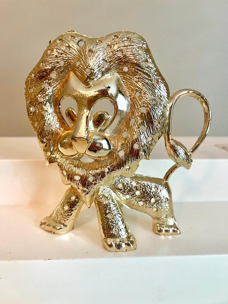 May include: A gold-colored lion figurine with a detailed mane and tail. The lion is standing on all fours, with a textured surface and small holes throughout. The figurine has a regal and decorative appearance.