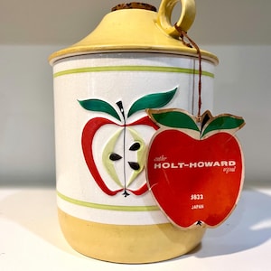May include: A ceramic canister with a yellow lid and base, featuring a white body with a green-lined apple motif. An apple-shaped paper tag is attached, printed with 'another HOLT-HOWARD original', '5832', and 'JAPAN' in white and black text.