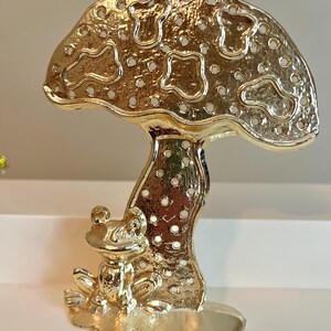 May include: A gold-colored metal mushroom-shaped figurine with a frog at its base. The mushroom cap has a perforated design with various shapes and small holes. The frog is detailed with visible eyes and limbs, and the figurine sits on a flat base.