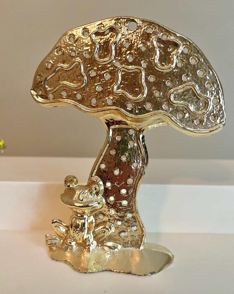 May include: A gold-colored metal mushroom-shaped figurine with a frog at its base. The mushroom cap has a perforated design with various shapes and small holes. The frog is detailed with visible eyes and limbs, and the figurine sits on a flat base.