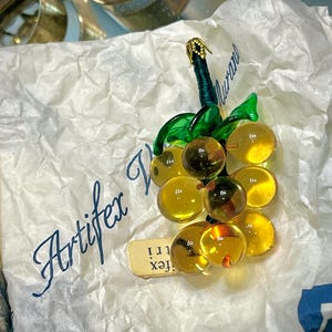 Op de afbeelding: Een glazen tros druiven met groene bladeren en een gouden steel. De druiven zijn doorschijnend amberkleurig. De tros ligt op wit tissuepapier met de tekst "Artifex Murano" in zwarte inkt.