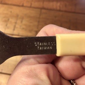 Op de afbeelding: Een close-up van een metalen bestek met een cr&egrave;mekleurige handgreep. Het bestek is gemarkeerd met "Stainless Taiwan".