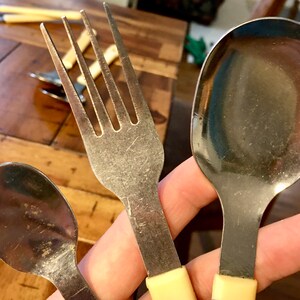 Op de afbeelding: Drie zilveren eetgerei met gele kunststof handvatten. Een vork, een lepel en een kleinere lepel.