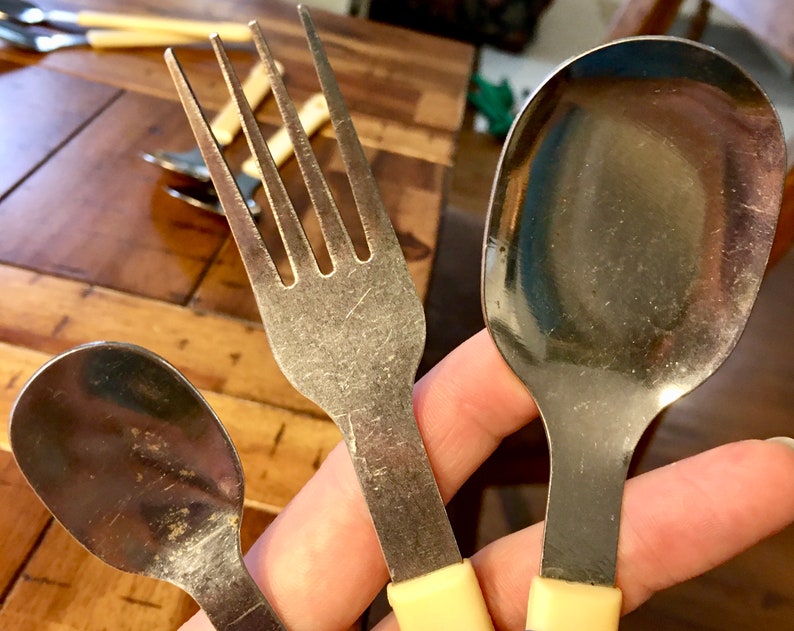 Op de afbeelding: Drie zilveren eetgerei met gele kunststof handvatten. Een vork, een lepel en een kleinere lepel.