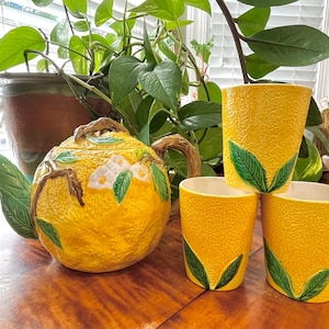 May include: A yellow ceramic teapot and three matching cups with a textured surface. The teapot has a branch handle and is decorated with white flowers and green leaves. The cups have green leaf accents.