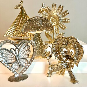 May include: A collection of decorative metal figurines in gold and silver tones. The figurines include a sailboat, mushroom, sunflower, butterfly in a heart, and a lion. The objects are displayed on a white surface.