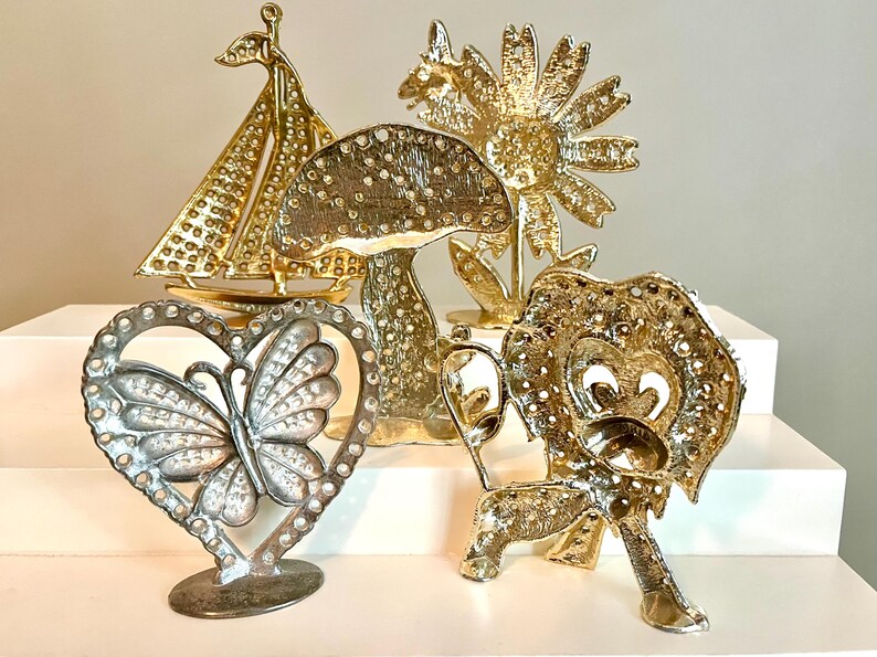 May include: A collection of decorative metal figurines in gold and silver tones. The figurines include a sailboat, mushroom, sunflower, butterfly in a heart, and a lion. The objects are displayed on a white surface.