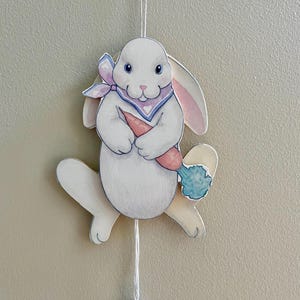 May include: A white wooden Easter bunny ornament with a pink and blue scarf, holding a carrot. The bunny has pink-lined ears and a blue and white carrot. A string and a lavender bead hang from the bottom.