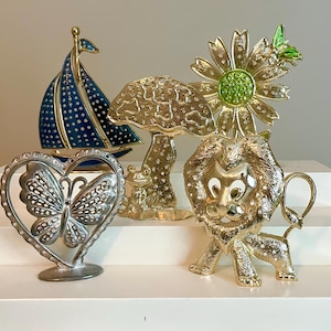 May include: A collection of vintage brooches and decorative objects. Includes a blue and gold sailboat, a gold mushroom with a frog, a gold flower with a green center and butterfly, a silver heart-shaped butterfly, and a gold lion.