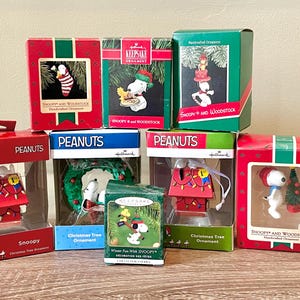 Op de afbeelding: Een verzameling Hallmark kerstversieringen met de personages Snoopy en Woodstock. De ornamenten zitten in hun originele dozen, met rode, groene en blauwe verpakkingen. De ornamenten tonen Snoopy, Woodstock en andere feestelijke ontwerpen.