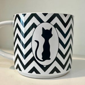 May include: White ceramic mug with a black and white chevron pattern. A black cat silhouette is centered on the mug within a white oval. The mug has a white handle.